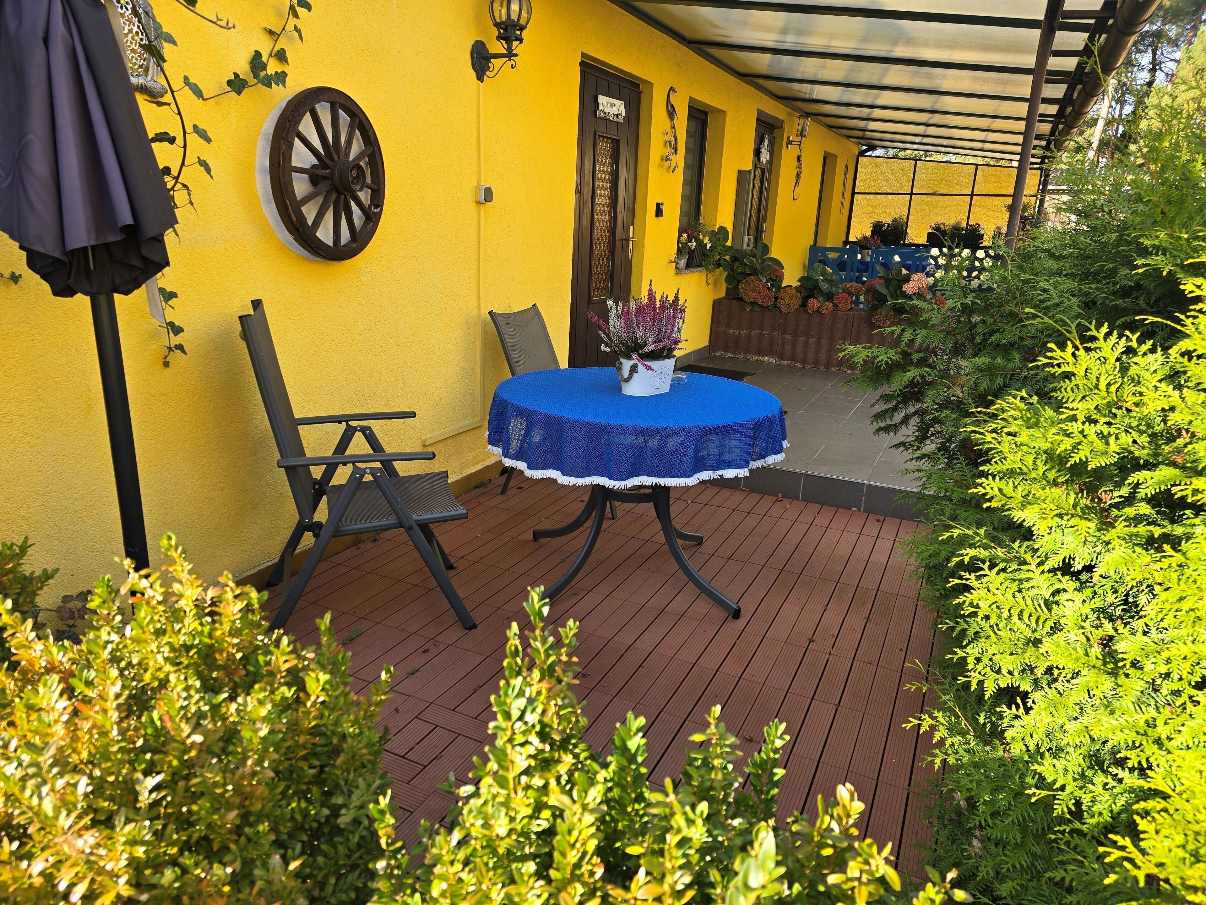 A covered patio with a table and chairs, surrounded by potted plants and a yellow wall.