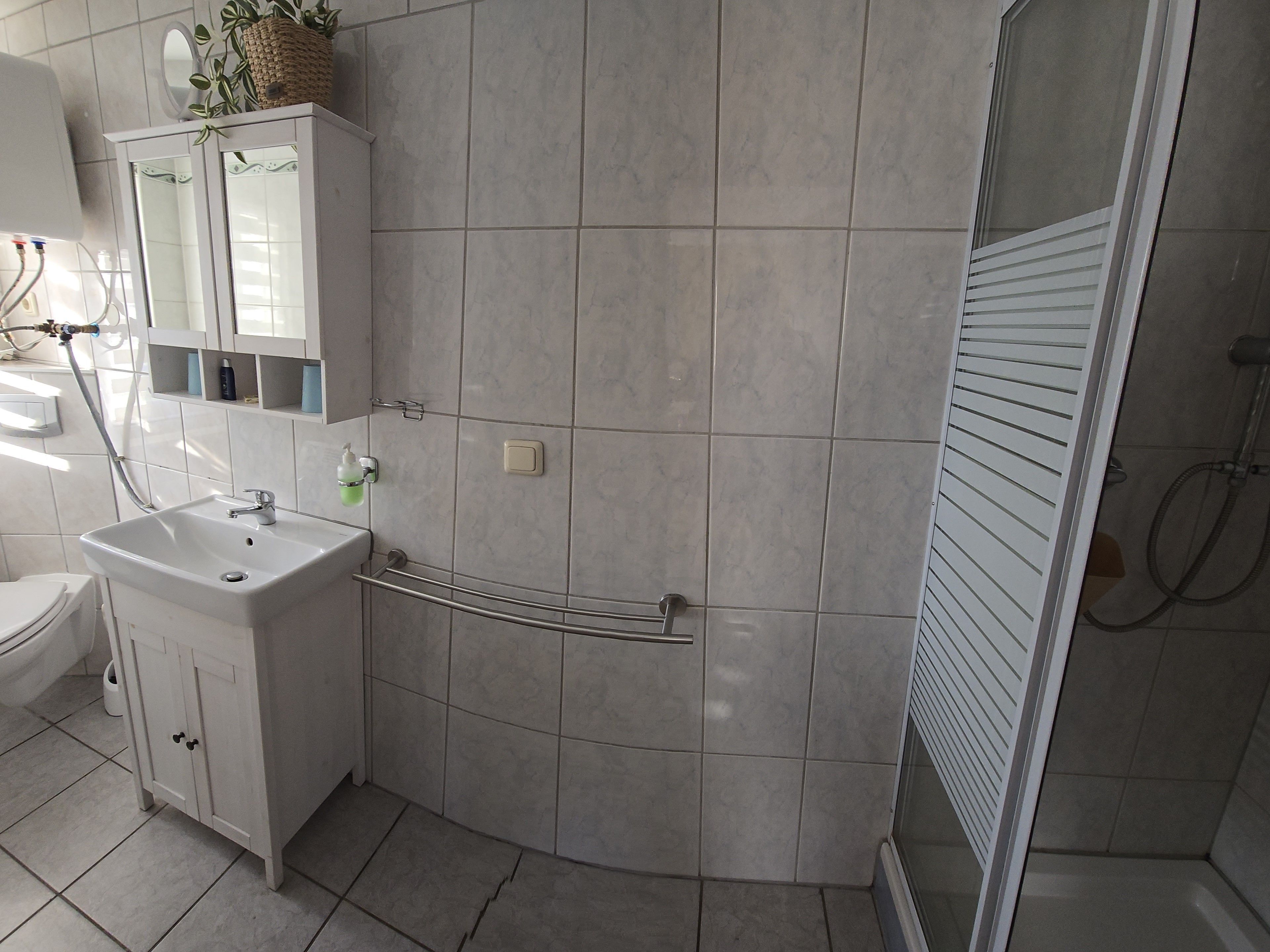 Et badeværelse med hvid vask, spejl og toilet. Der er et bruseområde med glasdør og et håndklædestativ.