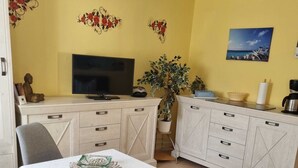 A compact living space with a dining table, a TV, a sofa, and a kitchen area with cabinets and a coffee machine. - Small Bungalow Wiesenperle - Small Bungalow meadow pearl (Trassenheide)