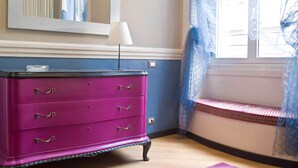A room with a pink dresser, a mirror, a lamp, and a window with blue curtains. - Hotel Colombo (Genoa)