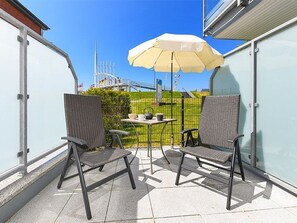 A balcony with two wicker chairs and a table, an umbrella, and a view of a bridge and greenery.