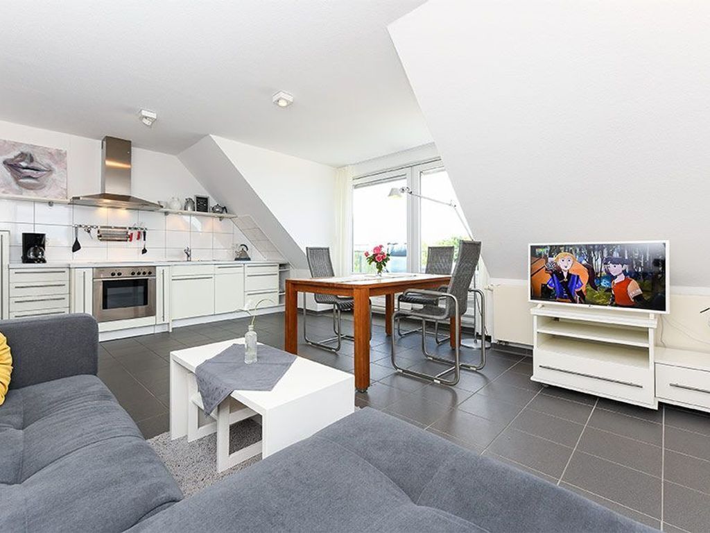 A modern living room with a grey sofa, a coffee table, and a TV on a stand. There is a dining area with a table and chairs, and a kitchen with white cabinets and a stove.