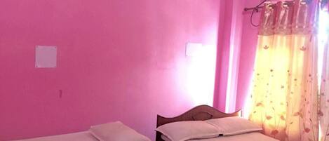 A room with pink walls, two single beds with white linens, a wooden headboard, a window with floral curtains, and a wall-mounted air conditioner.