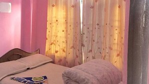 Un dormitorio con paredes rosas, una cama con una colcha, un almohadón y una mesita con un libro. Hay un equipo de aire acondicionado sobre la cama.
