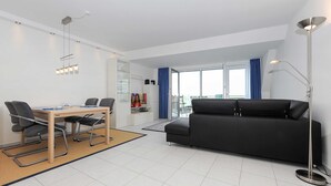 Uma sala de estar moderna com um sofá preto, uma mesa de jantar com cadeiras, uma janela grande com cortinas azuis e um abajur.