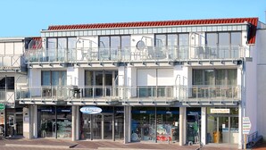 Um edifício de vários andares com telhado vermelho, apresentando uma loja no térreo com fachada de vidro e uma varanda com grades de metal.