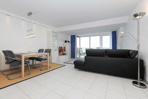 Ett modernt vardagsrum med en svart soffa, ett matbord med stolar, ett stort fönster med blå gardiner och en golvlampa.