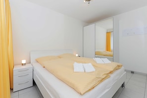 Uma cama de casal com manta bege e almofadas brancas, mesa de cabeceira com abajur, um espelho grande e uma parede branca com interruptor de luz.