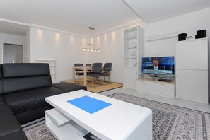 A modern living room with a black leather sofa, a white coffee table, a dining area with chairs, a television, and a cabinet with speakers. - Vacation apartment Benser Nest - Residence Hafenidyll (Esens)