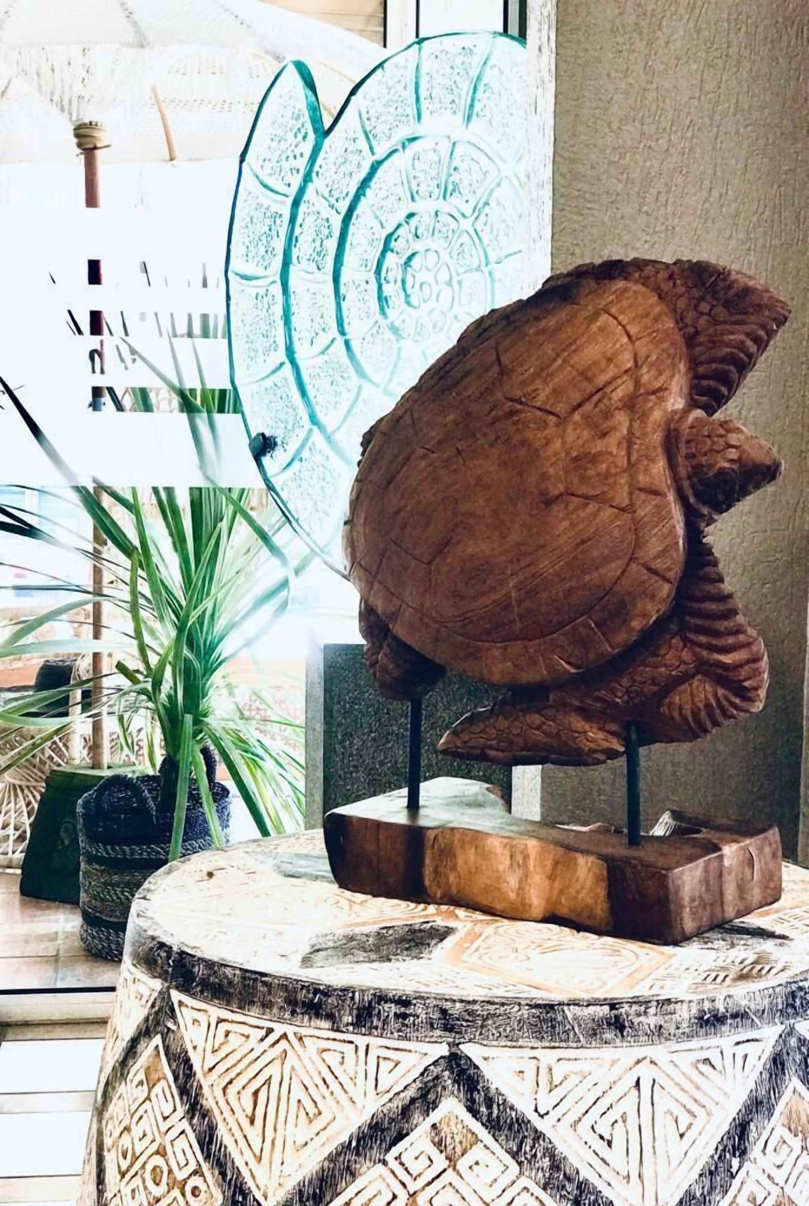 A wooden turtle sculpture on a round table with a patterned base, surrounded by indoor plants and a large window with a view of buildings.