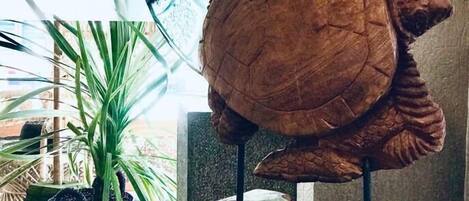 A wooden turtle sculpture on a round table with a patterned base, surrounded by indoor plants and a large window with a view of buildings.
