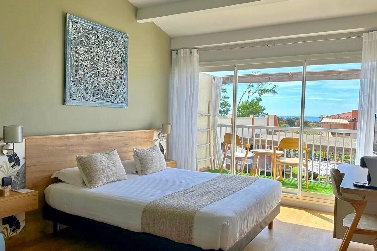 A bedroom with a large bed, a desk with a chair, a balcony with chairs, and a view of the outside.