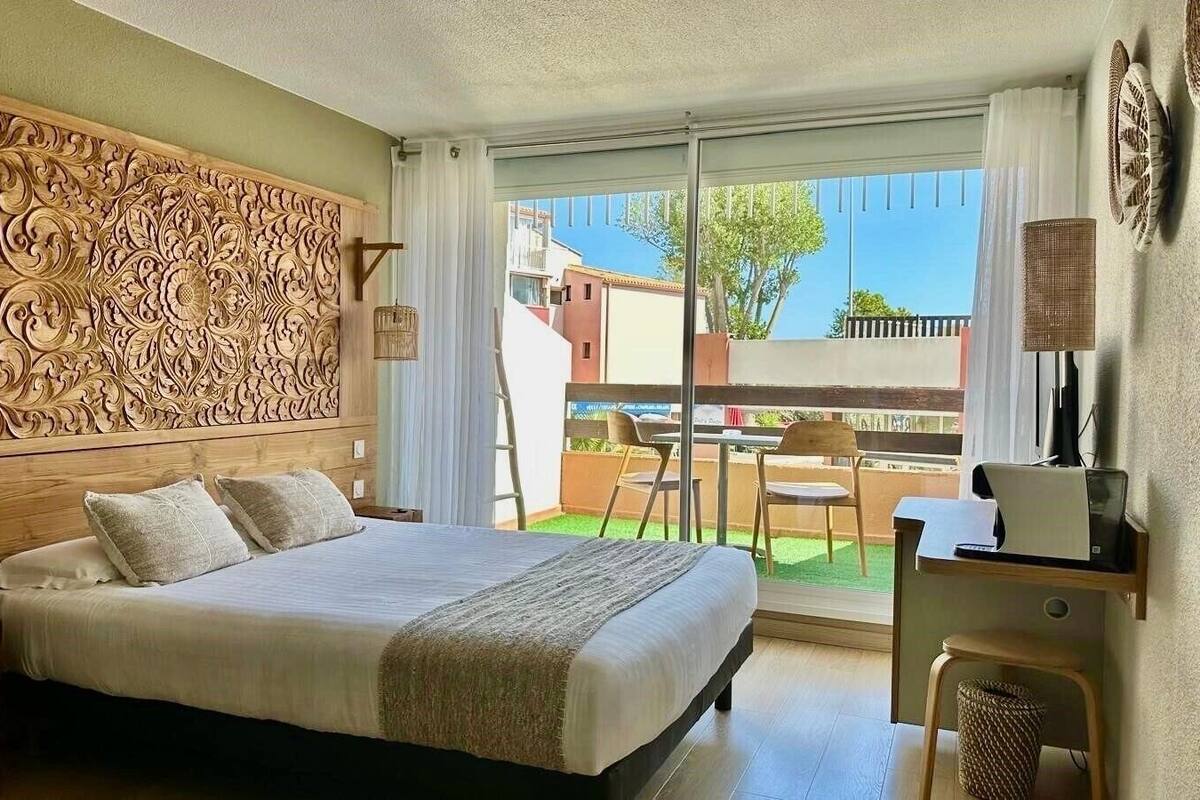 A bedroom with a large bed, a wooden headboard with intricate carvings, a desk with a chair, and a view of the outside balcony.