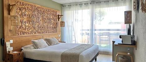 Une chambre à coucher avec un grand lit, une tête de lit en bois ornée de motifs complexes, une table de chevet, une lampe et une vue sur l’extérieur grâce à une grande fenêtre.