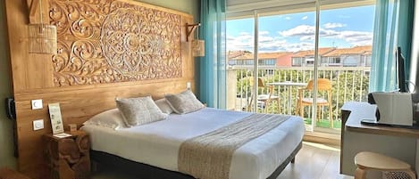 Een hotelkamer met een groot bed, een houten hoofdbord met ingewikkelde decoraties, een klein houten nachtkastje en een balkon met buitenmeubilair.