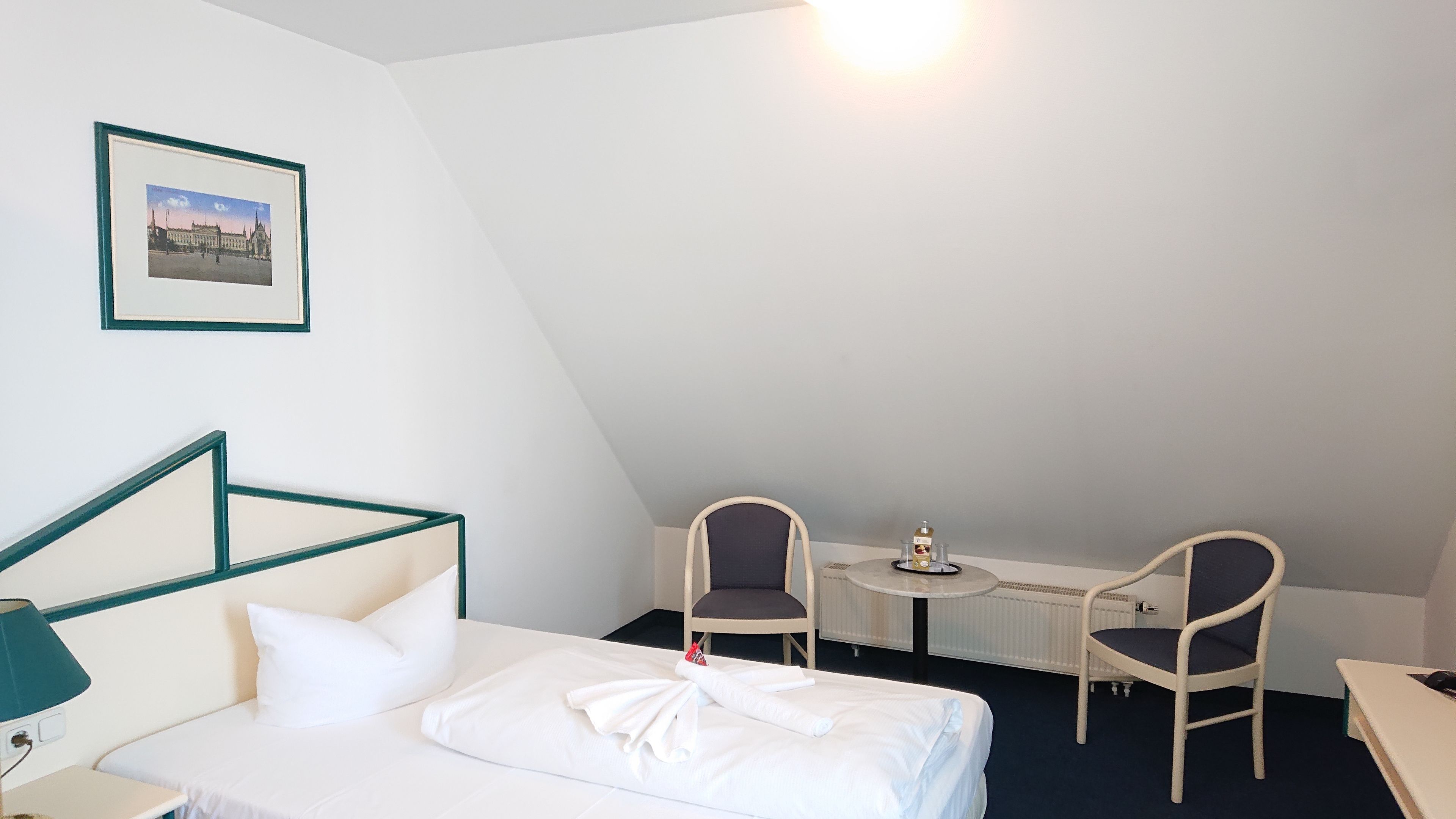 Ett hotellrum med en säng, ett skrivbord, två stolar, ett litet bord med en flaska och glas, och en inramad tavla på väggen.