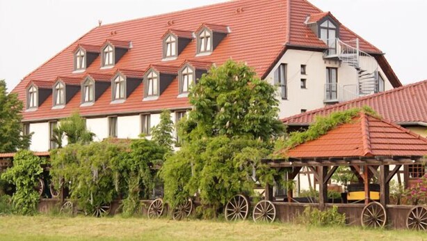 A large building with a red-tiled roof, multiple windows, and a covered area with wooden beams and wheels. - Hotel 3 Linden (Leipzig)