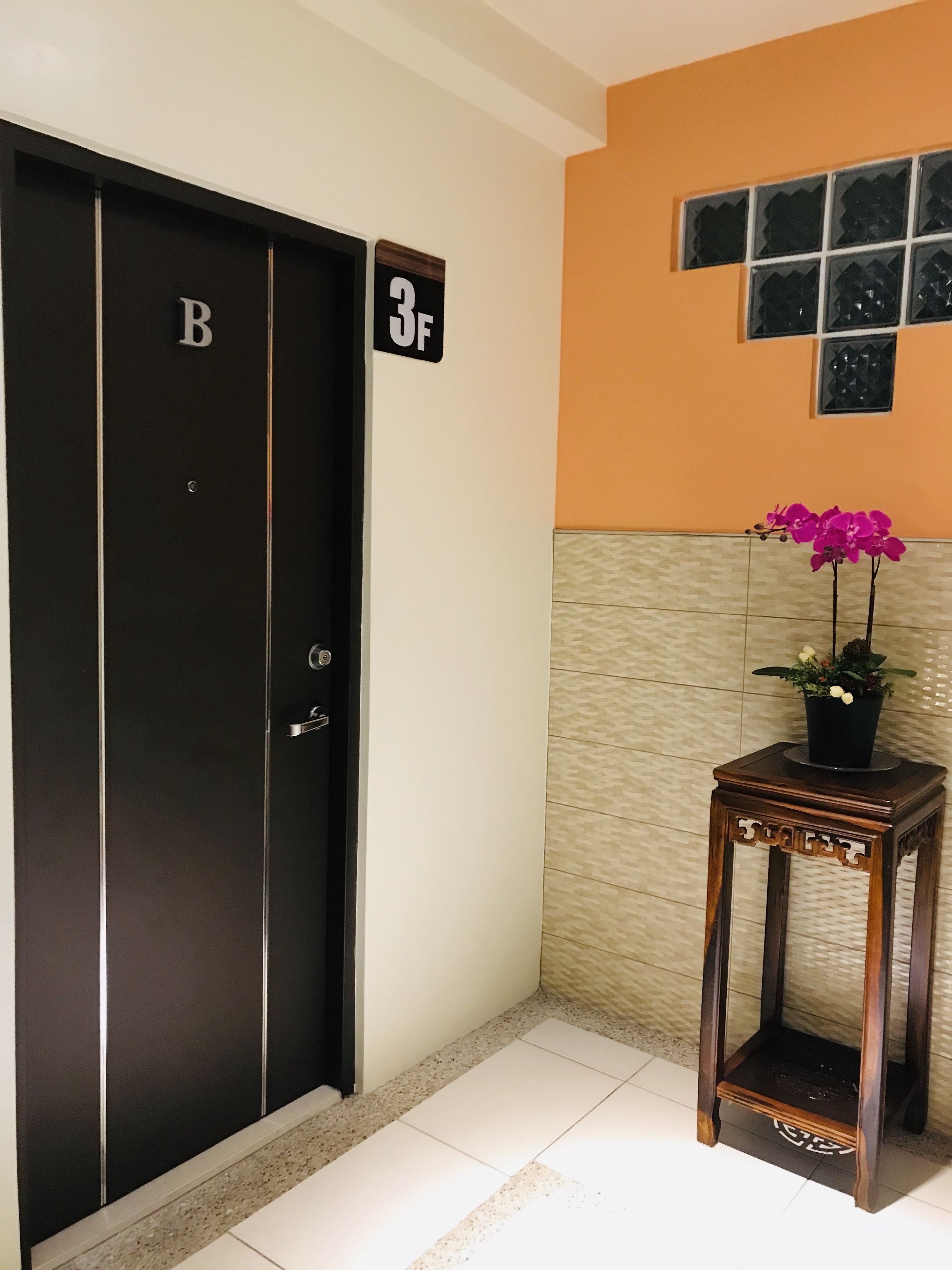 Hotelkamer met een zwarte deur waarop 'B' en '3F' staan, een klein houten tafeltje met een potorkidee en een betegelde vloer.