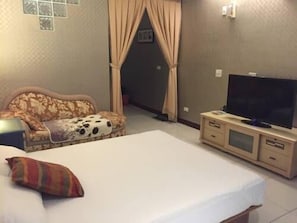 Quarto de hotel com cama, televisão sobre suporte de madeira, sofá com capa estampada e porta com cortinas.