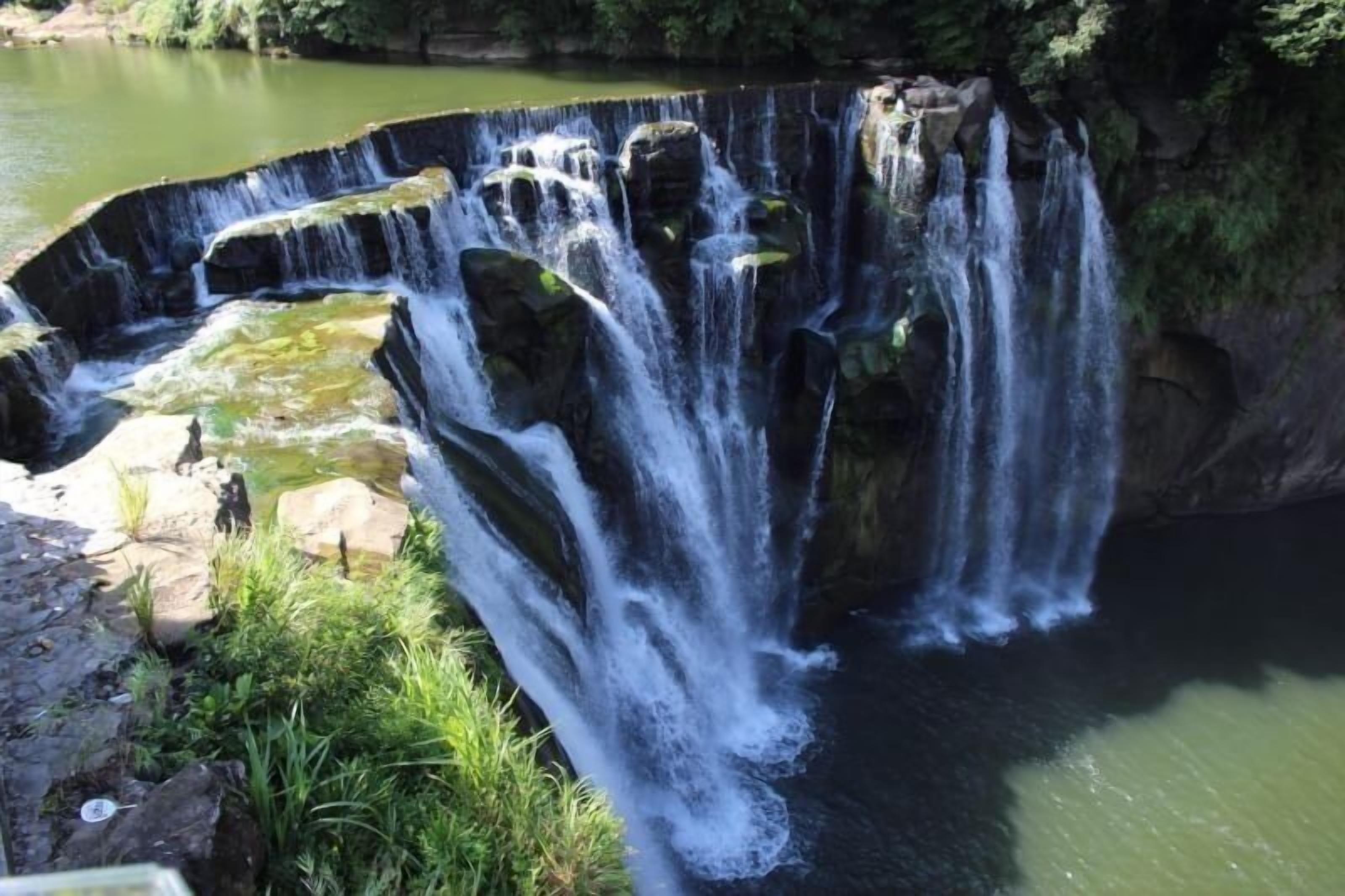 A waterfall cascading over a series of rocky steps into a calm body of water, surrounded by greenery.