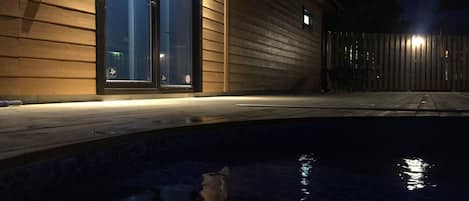 A nighttime view of a swimming pool with a wooden deck and a building with glass doors.