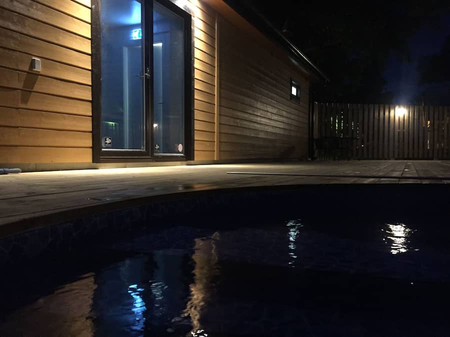 A nighttime view of a swimming pool with a wooden deck and a building with glass doors.