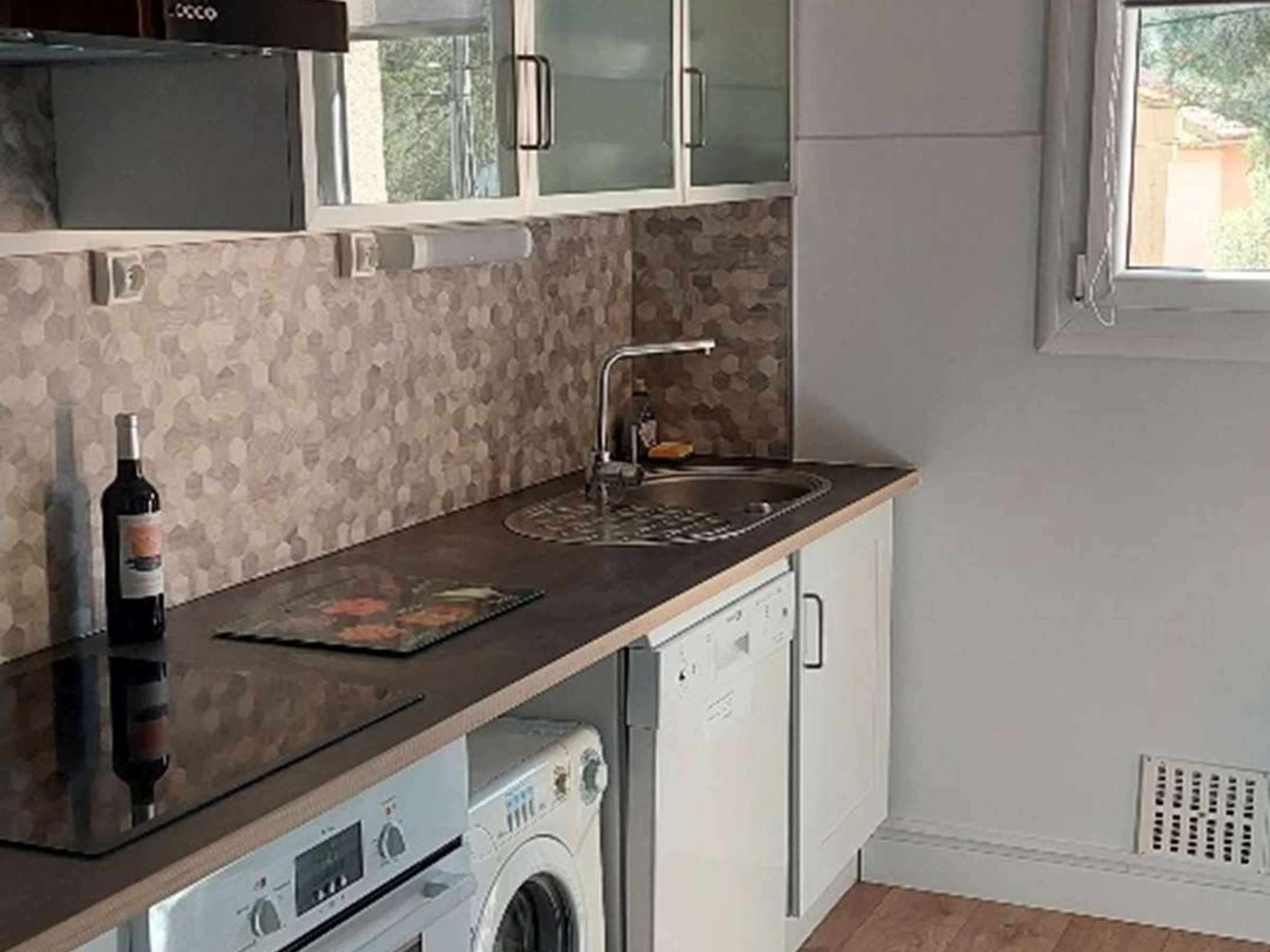 Een compacte keuken met een geïntegreerde wasmachine, een spoelbak en een fles wijn op het aanrecht.