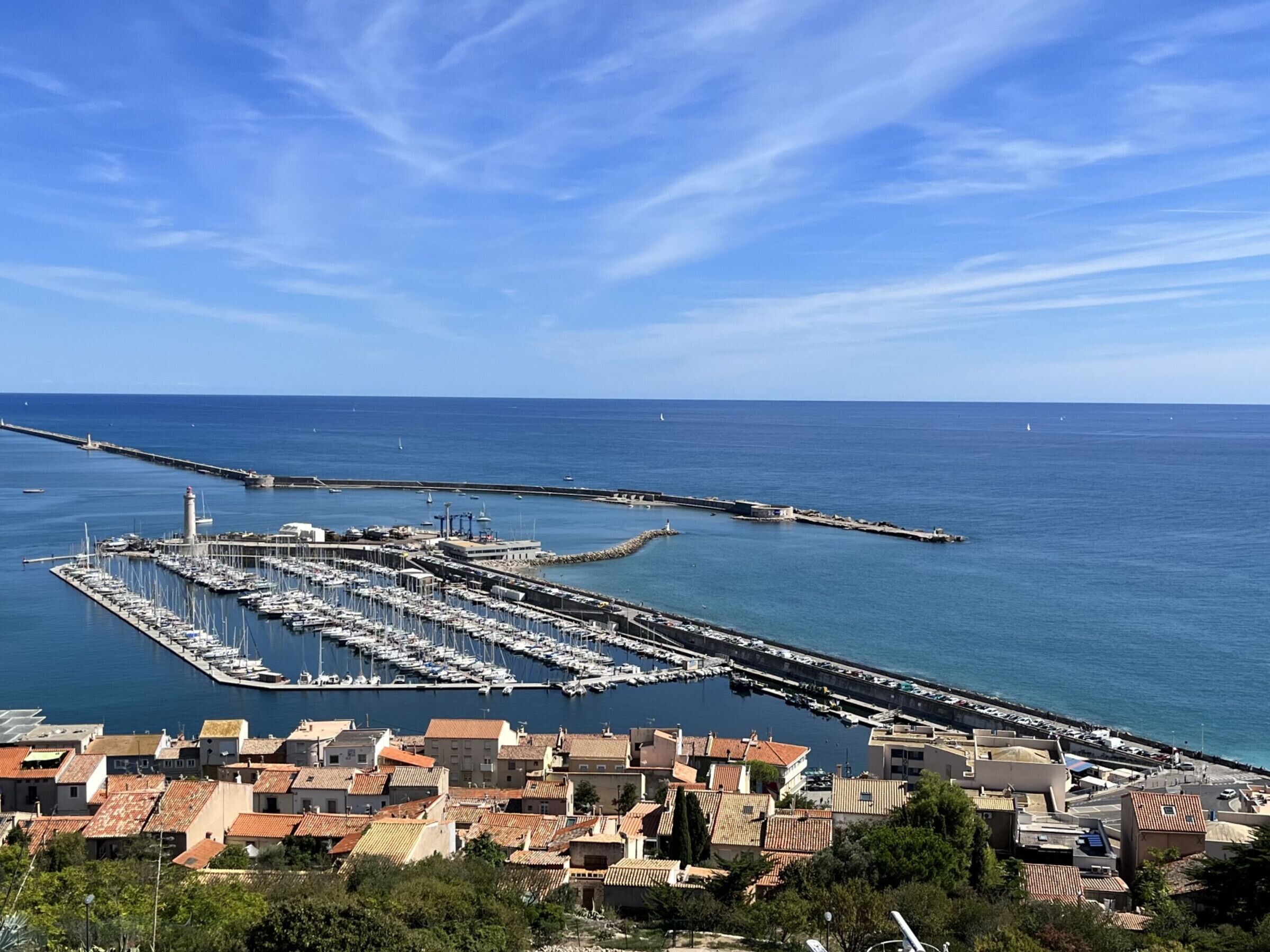 Un port de plaisance situé sur la côte, avec des bateaux amarrés, un phare et une jetée qui s’avance dans la mer.