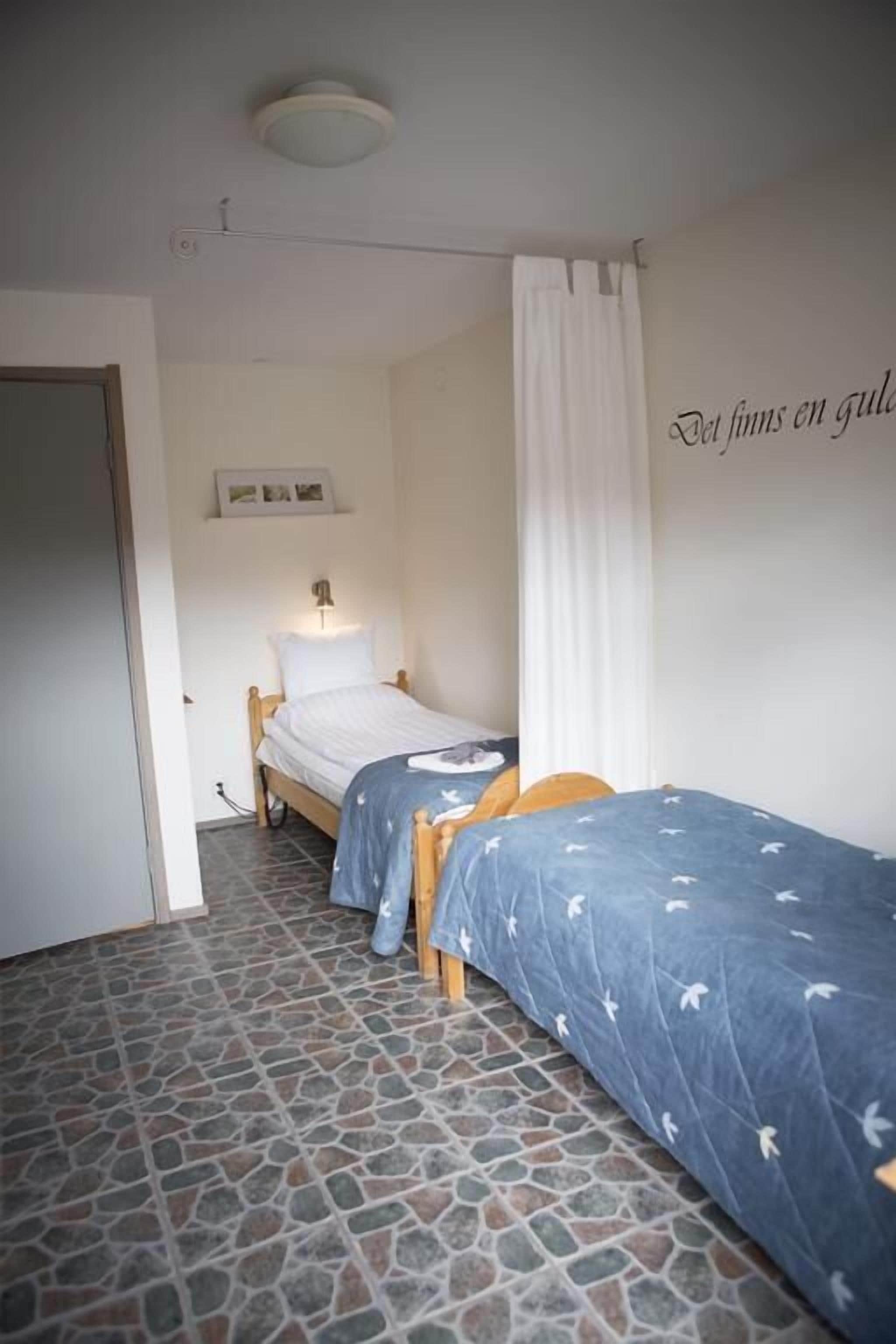 Una habitación pequeña y acogedora con dos camas individuales, piso de baldosas de piedra, una cortina blanca y una pared con una frase en francés.