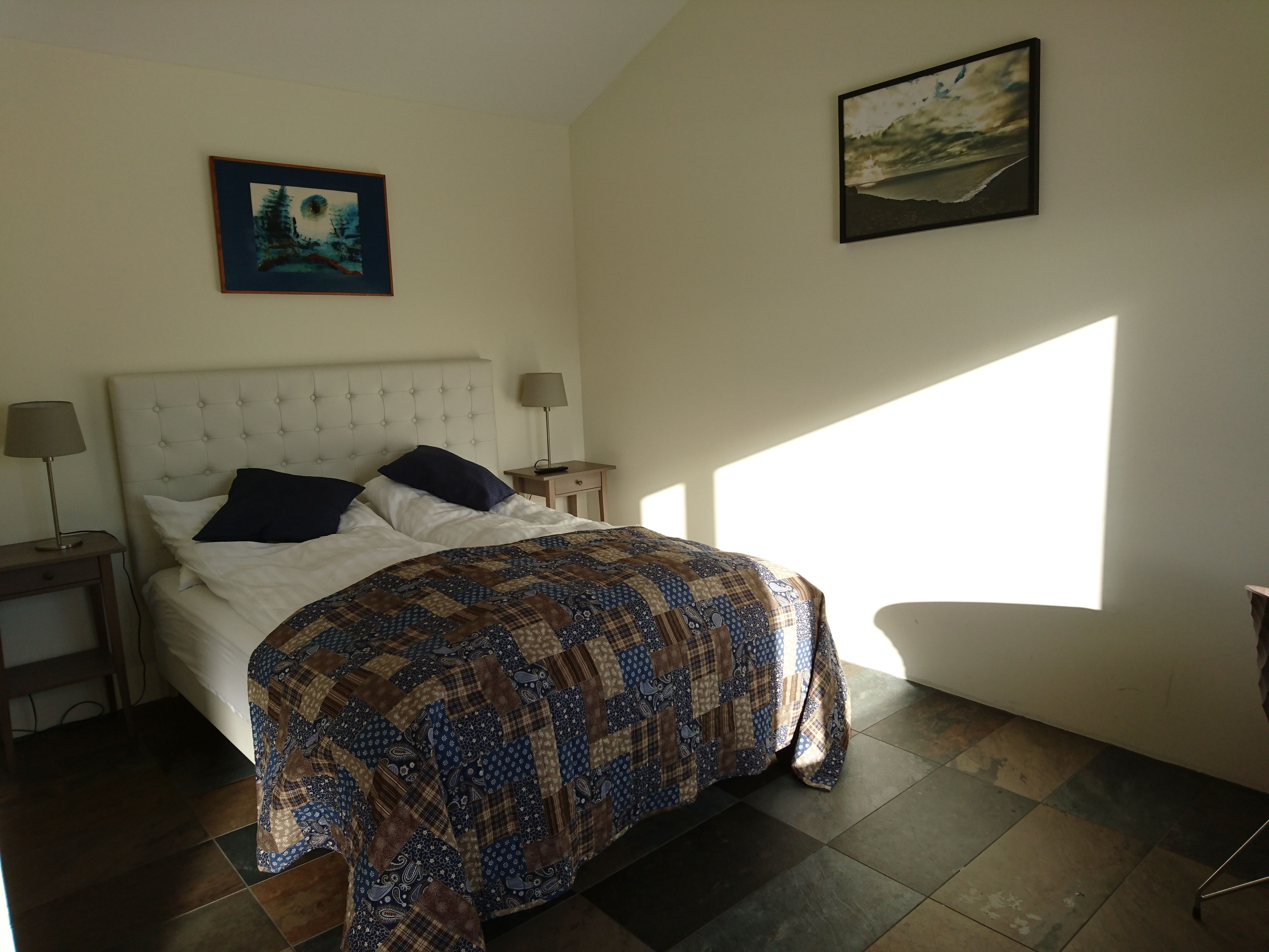 Un dormitorio con una cama, dos mesitas de noche, una lámpara y dos cuadros colgados en la pared.