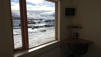 Habitación con amplia ventana que da a un paisaje nevado, un radiador, una mesita con un hervidor y un televisor montado en la pared.