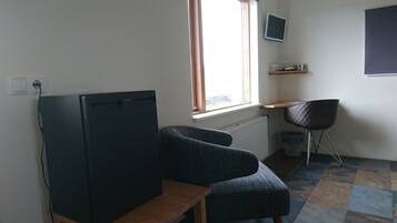 Um quarto pequeno com televisão, uma cadeira, uma janela e um radiador.