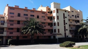 A multi-story residential building with a uniform facade, balconies, and a parking area in front.