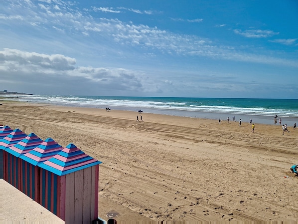 Une plage où l’on voit des gens se promener et jouer, un rivage sablonneux et un bâtiment doté d’auvents à rayures.