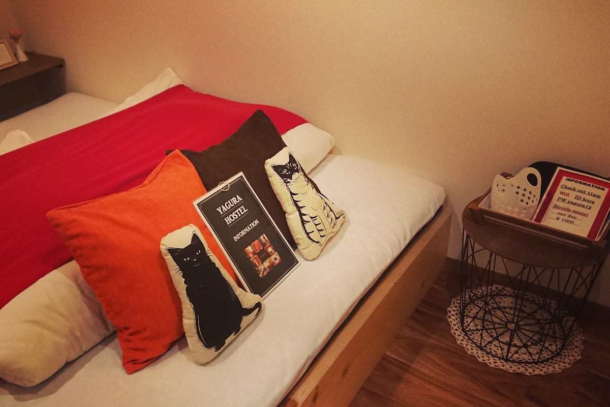 Ein Bett mit roter Decke, ein Kopfkissen mit Katzenmotiv in Schwarz und ein Kopfkissen mit Katzenmotiv in Weiß. Ein kleiner Tisch mit einem Schild und einem dekorativen Gegenstand.