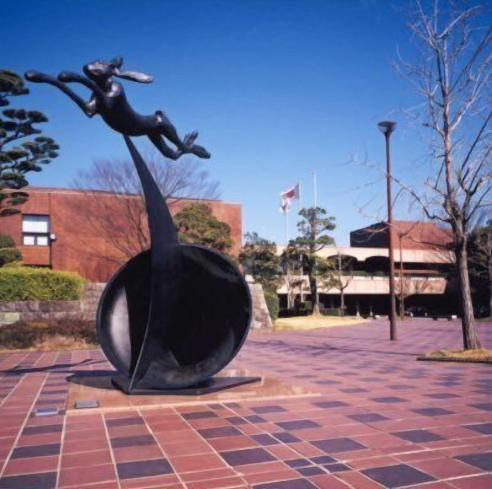 A sculpture of a figure in a dynamic pose, set on a tiled plaza with a building in the background.