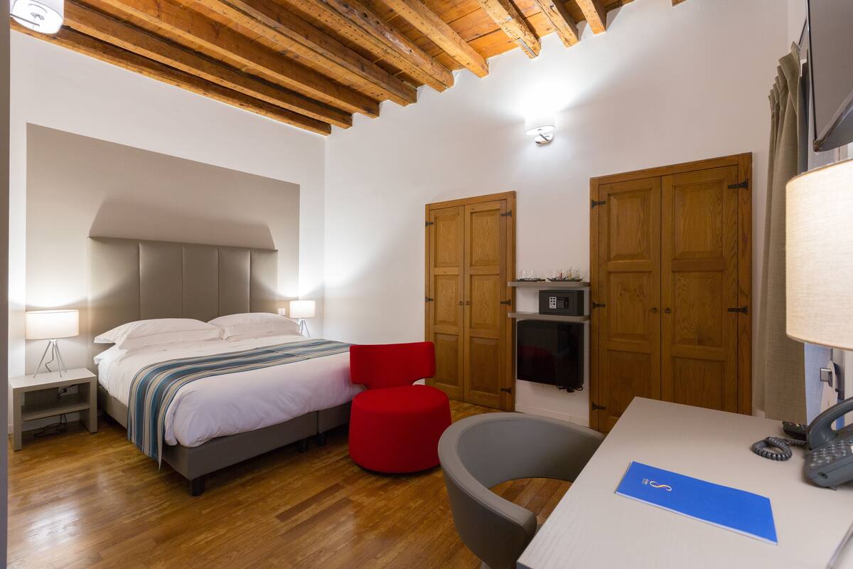 A hotel room with a bed, a desk with a telephone, a red chair, and wooden doors.