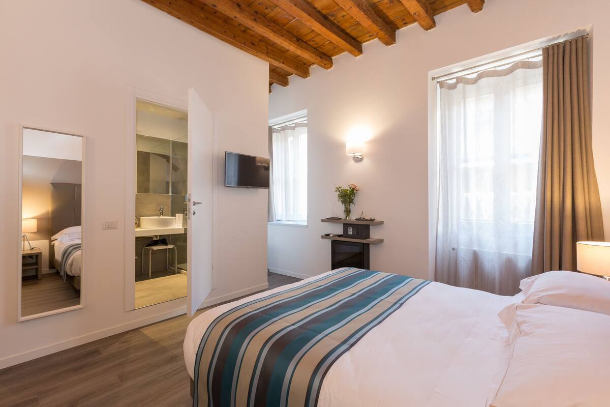 A bedroom with a bed, a TV, a mirror, a bathroom, and a window with curtains.