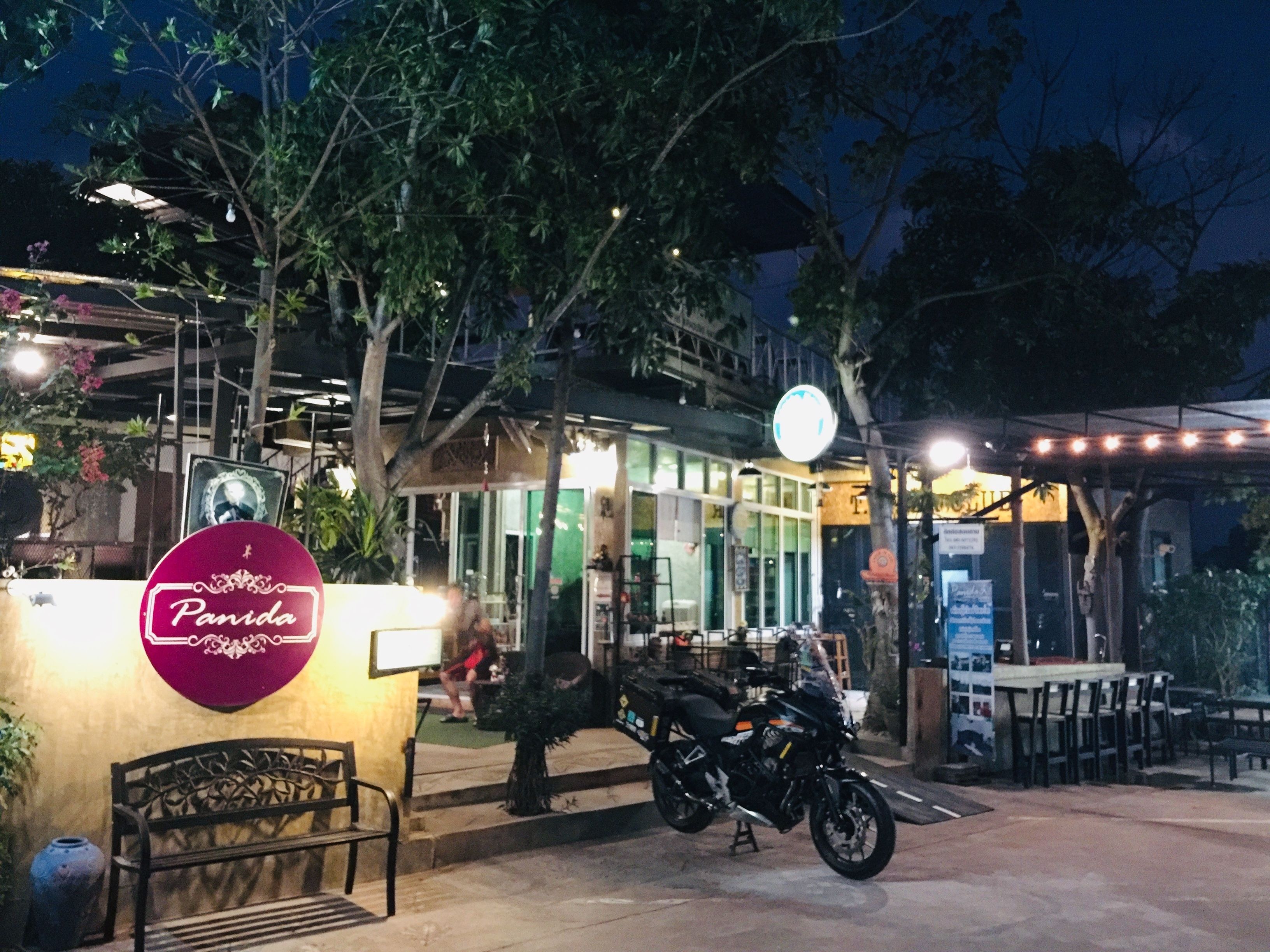 Un espacio de estar al aire libre con una motocicleta estacionada cerca, un cartel que dice "Panida" y un edificio con puertas de cristal al fondo.
