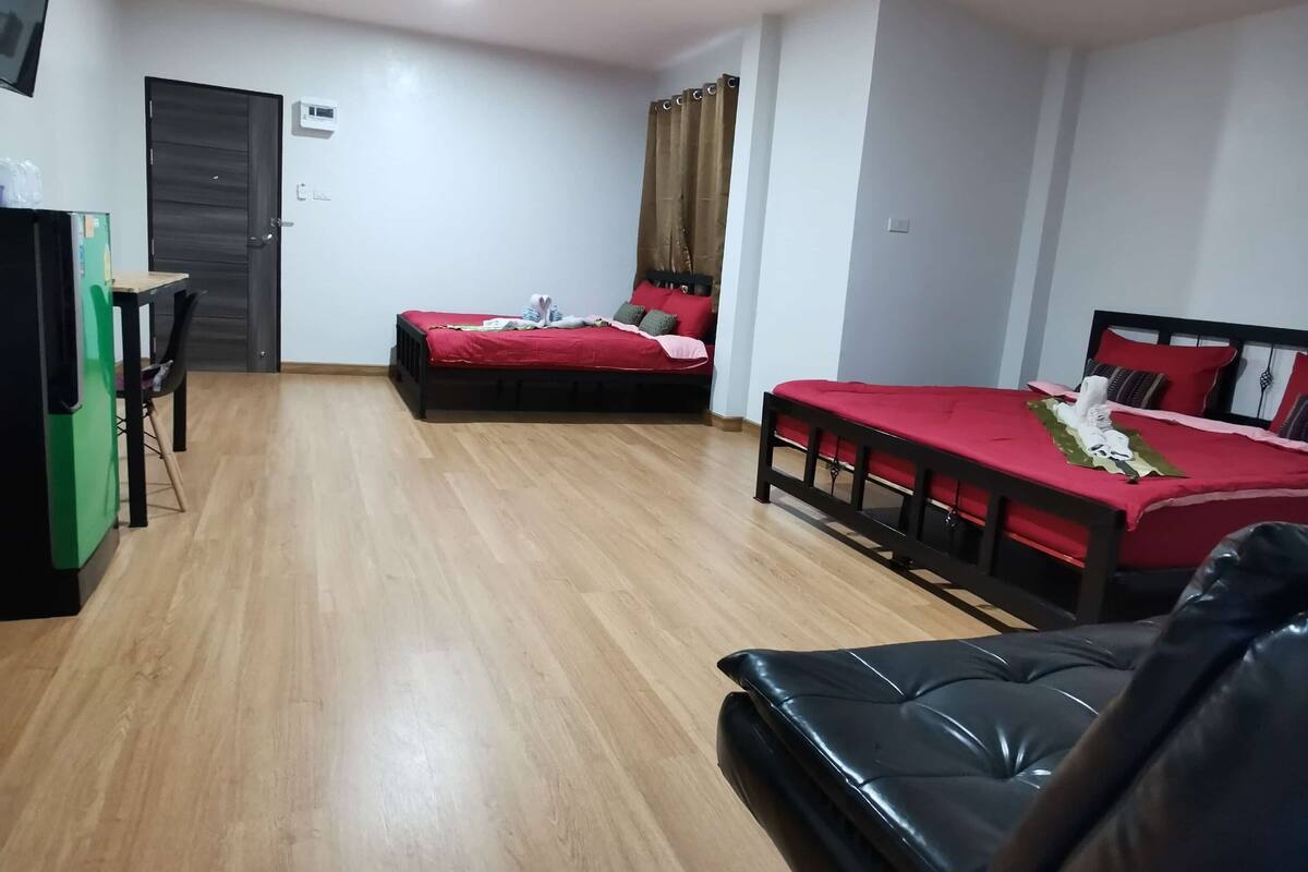 Una habitación con suelo de madera, un sofá de cuero negro, dos camas individuales con ropa de cama roja, una mesita con sillas y una puerta con unidad de aire acondicionado.