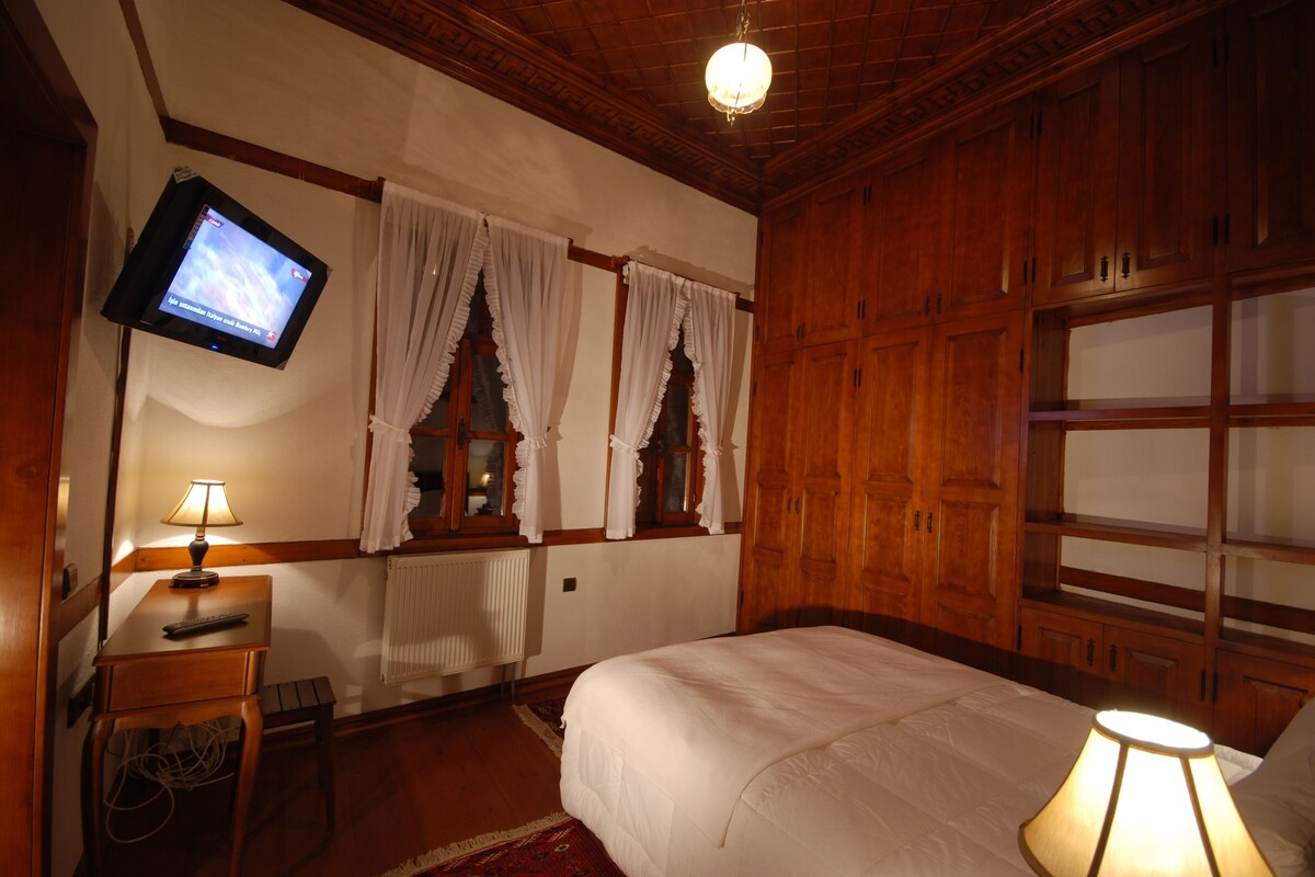 Ein Schlafzimmer mit Holzboden, einem an der Wand montierten Fernseher, einem Bett mit weißer Bettwäsche, einem Nachttisch mit Lampe und einem Fenster mit Vorhängen.
