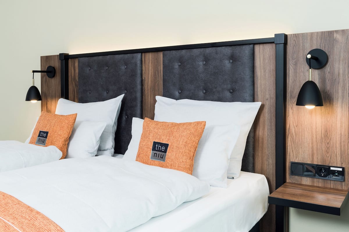 A neatly made bed with white and orange bedding, a dark headboard, and two wall-mounted reading lamps.
