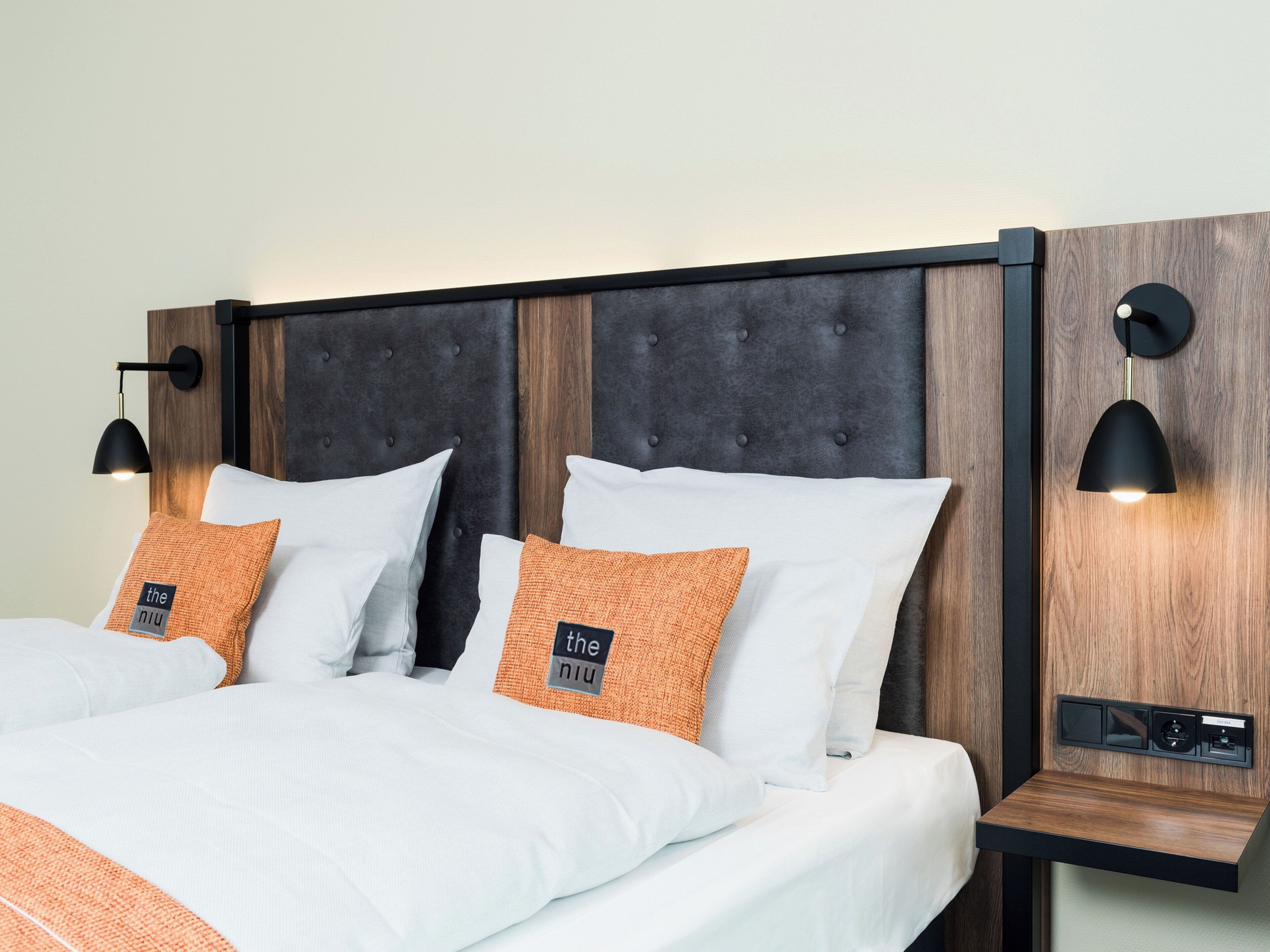 A neatly made bed with white and orange bedding, a dark headboard, and two wall-mounted reading lamps.