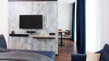 Una habitación de hotel moderna con una televisión de pantalla plana montada en una pared de madera decorativa, una zona de comedor con mesa y sillas, y un ventanal con cortinas azules.