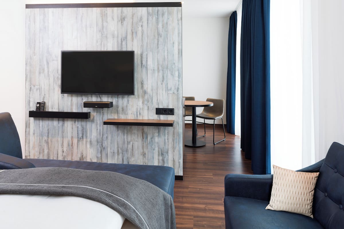 A modern hotel room with a flat-screen TV mounted on a wooden accent wall, a dining area with a table and chairs, and a large window with blue curtains.