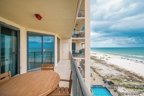 A balcony with a view of the beach, a pool, and a dining area with a table and chairs.