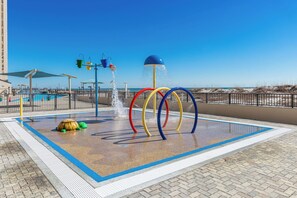 Un espace de jeux aquatiques avec des anneaux colorés et une fontaine, entouré d’une piscine et offrant une vue sur la plage.