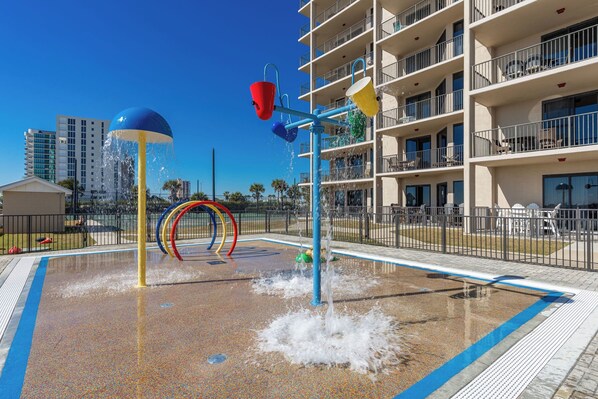 A water play area with a pool, surrounded by a fence, and a multi-story building in the background. - Book Today! - Phoenix X - 8th Floor 3BR Condo! (Orange Beach)
