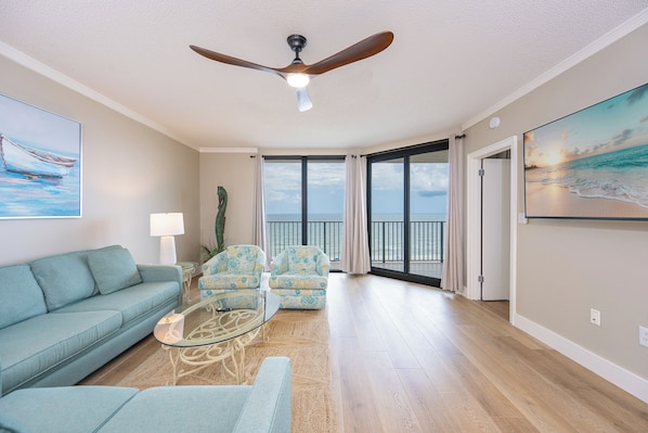 A spacious living room with a ceiling fan, a sofa, armchairs, a glass coffee table, and large windows offering a view of the ocean.