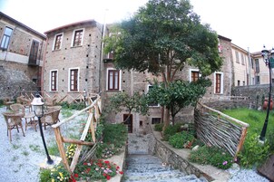 Un edificio de piedra rústico con una escalera de madera que conduce a una puerta, rodeado de abundante vegetación y un pequeño jardín con flores.